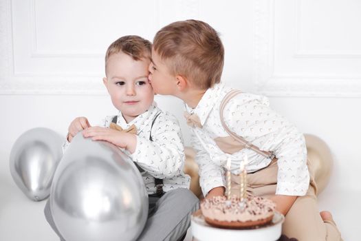 Two boys celebrating birthday, children have a B-day party. Birthday cake with candles and balloons. Happy kids, celebration, white minimalist interior