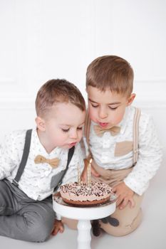 Two boys celebrating birthday, children have a B-day party. Kids blowing candles on the birthday cake. Celebration, white minimalist interior