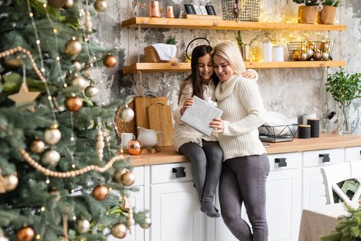 Beautiful mother and daughter have fun, hug and kiss at home near the Christmas tree in a white interior. Family happiness, holiday, joy, vacation, games with a woman. New Year's preparations.