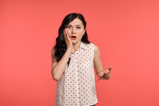Studio shot of a good-looking girl teenager, wearing casual white polka dot blouse. Little brunette female is looking shoked, posing over a pink background. People and sincere emotions.