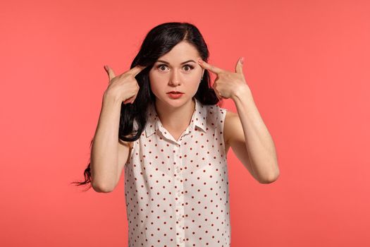 Studio shot of an amazing teeny girl looking shocked, wearing casual white polka dot blouse. Little brunette female put her index fingers to her head posing over a pink background. People and sincere emotions.