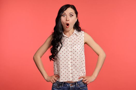 Studio shot of a graceful girl teenager, wearing casual white polka dot blouse. Little brunette female is looking wondered, posing over a pink background. People and sincere emotions.