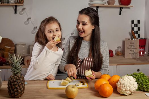 Happy loving family are cooking together. Cute mother and her little princess are doing a fruit cutting and tasting an apple at the kitchen, against a white wall with shelves and bulbs on it. Homemade food and little helper.