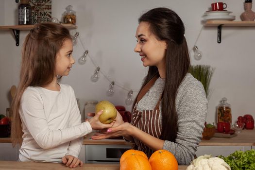 Happy loving family are cooking together. Loving mum and her child are doing a fruit cutting and sharing with apple at the kitchen, against a white wall with shelves and bulbs on it. Homemade food and little helper.