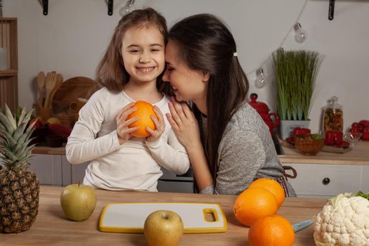 Happy loving family are cooking together. Loving mother and her daughter are doing a fruit cutting and hugging at the kitchen, against a white wall with shelves and bulbs on it. Homemade food and little helper.