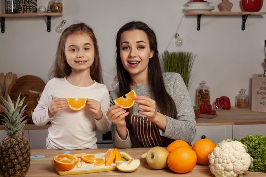 Happy loving family are cooking together. Attractive mom and her kid are doing a fruit cutting, smiling and holding an orange at the kitchen, against a white wall with shelves and bulbs on it. Homemade food and little helper.