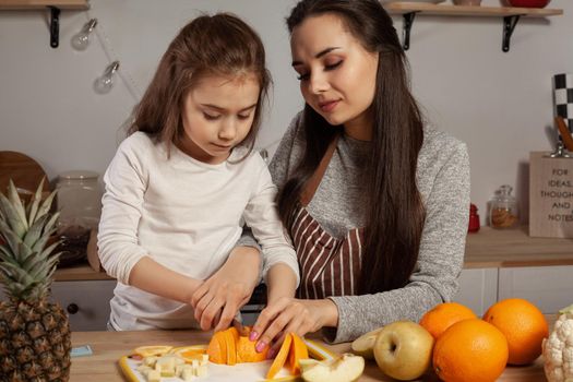 Happy loving family are cooking together. Elegant mother and her little princess are doing a fruit cutting and having fun at the kitchen, against a white wall with shelves and bulbs on it. Homemade food and little helper.
