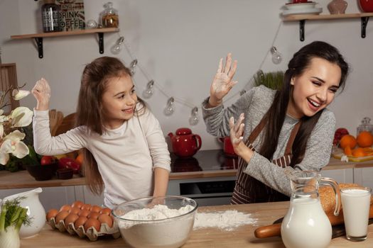 Happy loving family are preparing pastries together. Lovely mommy and her little girl are throwing a flour in each other and having fun at the kitchen, against a white wall with shelves and bulbs on it. Homemade food and little helper.