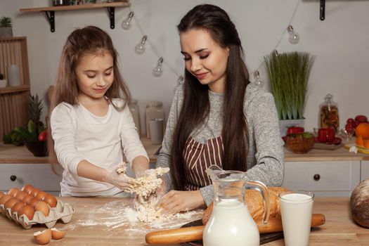 Happy loving family are preparing pastries together. Attractive mum and her child are kneading a dough and having fun at the kitchen, against a white wall with shelves and bulbs on it. Homemade food and little helper.