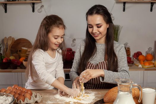 Happy loving family are preparing pastries together. Alluring mom and her kid are adding an egg to the dough and having fun at the kitchen, against a white wall with shelves and bulbs on it. Homemade food and little helper.