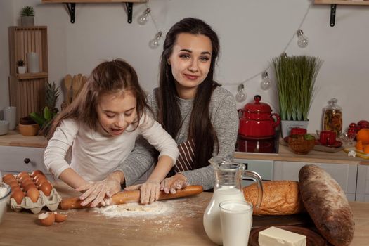 Happy loving family are preparing pastries together. Beautiful mother and her little princess are rolling a dough and having fun at the kitchen, against a white wall with shelves and bulbs on it. Homemade food and little helper.