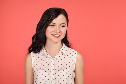 Close-up studio shot of a gorgeous teeny girl, wearing casual white polka dot blouse. Little brunette female is smiling and looking away posing over a pink background. People and sincere emotions.