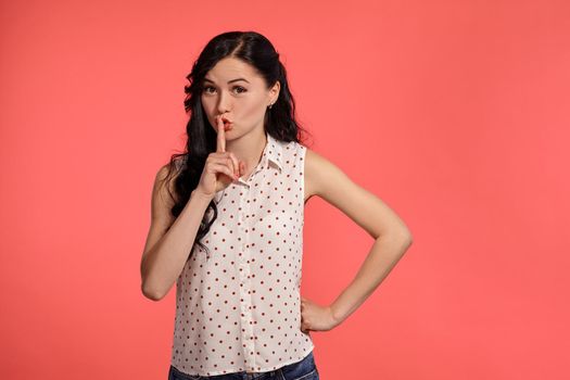 Studio shot of a gorgeous teen lady, wearing casual white polka dot blouse. Little brunette female is showing quiet sign while posing over a pink background. People and sincere emotions.