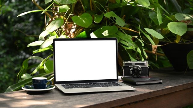 Computer laptop with white screen, coffee cup and camera on wooden table in the green garden.