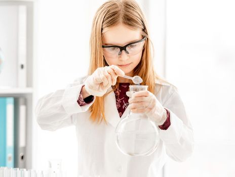 Smart girl doing scientific chemistry experiment wearing protection glasses, holding bottle and measuring ingridients. Schoolgirl with chemical equipment on school lesson