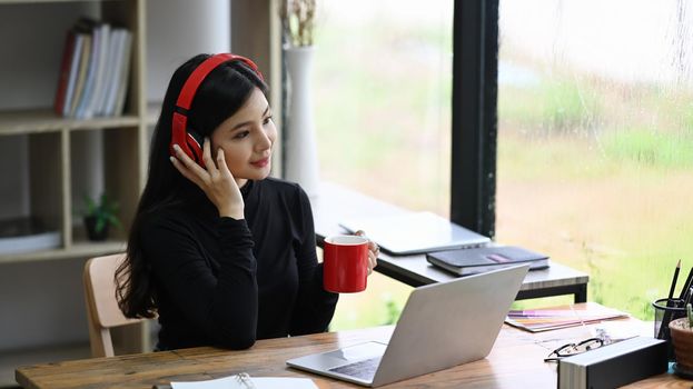 Attractive young woman designer wearing headphone and drinking coffee in office.