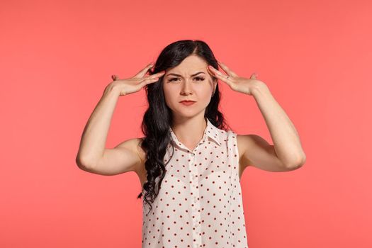 Studio shot of an alluring adolescent girl looking unhappy, wearing casual white polka dot blouse. Little brunette female acting like she has got an headache, posing over a pink background. People and sincere emotions.