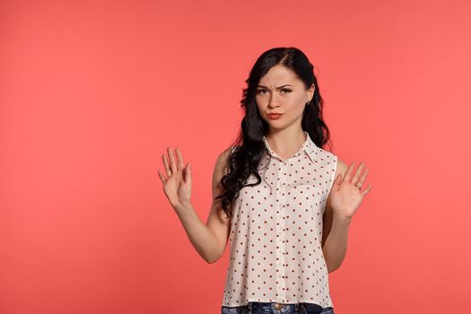 Studio shot of a lovely adolescent girl looking at the camera, wearing casual white polka dot blouse and denim short shorts. Little brunette female acting like denies something posing over a pink background. People and sincere emotions.