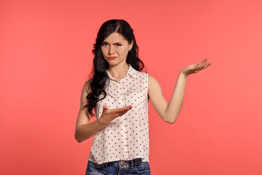 Studio shot of a lovely little woman, wearing casual white polka dot blouse and denim short shorts. Little brunette female looking upset, posing over a pink background. People and sincere emotions.