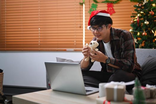 Smiling young man showing teddy bear during video call with his girlfriend at Christmas time.