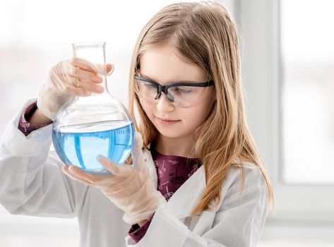Smart girl doing scientific chemistry experiment wearing protection glasses, holding bottle and measuring blue liquid. Schoolgirl with chemical equipment on school lesson