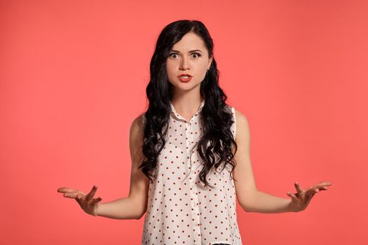 Studio shot of an attractive little woman looking angry, wearing casual white polka dot blouse. Little brunette female posing over a pink background. People and sincere emotions.