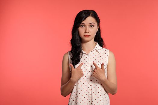 Studio shot of an amazing girl teenager looking surprised, wearing casual white polka dot blouse. Little brunette female is posing over a pink background. People and sincere emotions.