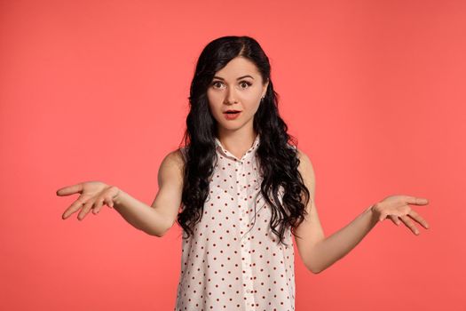 Studio shot of an attractive girl teenager looking confused, wearing casual white polka dot blouse. Little brunette female is posing over a pink background. People and sincere emotions.