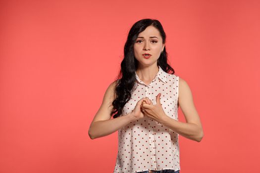 Studio shot of a pretty adolescent girl looking confused, wearing casual white polka dot blouse. Little brunette posing over a pink background. People and sincere emotions.