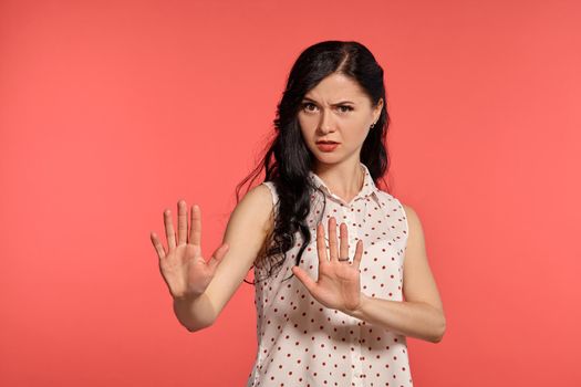 Studio shot of a charming teen lady, wearing casual white polka dot blouse. Little brunette female feeling unhappy acting like she is stopping someone, posing over a pink background. People and sincere emotions.