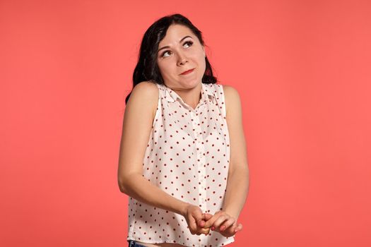 Studio shot of an alluring girl teenager looking shy, wearing casual white polka dot blouse. Little brunette female posing over a pink background. People and sincere emotions.