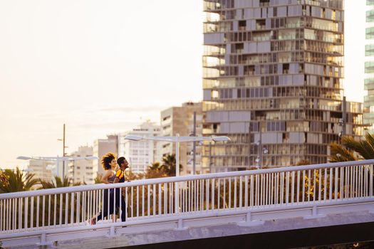 beautiful latin woman and happy black man jogging together at sunset in the city bridge with buildings in the background. urban sports workout in couple and healthy lifestyle concept