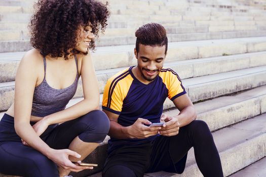 portrait of an athletic interracial couple having fun with mobile phone on the stairs during a rest in the workout, technology in sport and healthy lifestyle concept