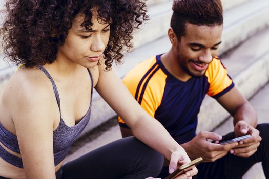 portrait of a athletic latin woman and black man using their mobile phones on the stairs during a rest in the workout, technology in sport and healthy lifestyle concept