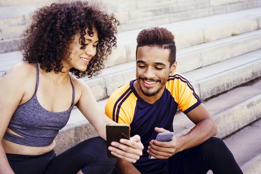 happy athletic latin woman shows the sports app in her mobile phone to a black sporty man on the stairs during a rest in the workout, technology in sport and healthy lifestyle concept