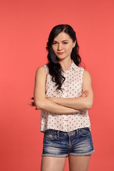 Studio shot of a charming smiling teeny girl, wearing casual white polka dot blouse and denim short shorts. Little brunette crossed her hands, posing over a pink background. People and sincere emotions.