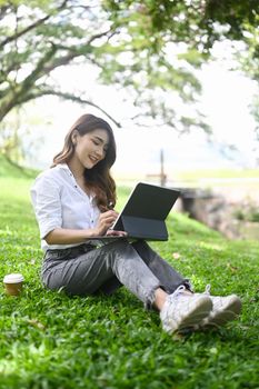 Young female freelancer sitting on grass in public park and working with computer tablet.