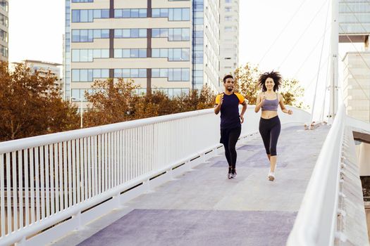 handsome black man and happy latin woman running together in a city bridge with buildings in the background. urban sports workout in couple and healthy lifestyle concept