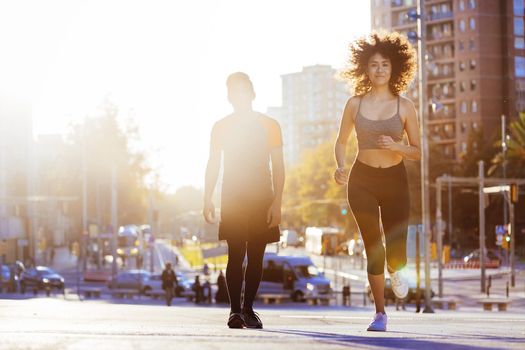 beautiful latin woman running at sunset in the city street with buildings and the silhouette of a sports man in the background. urban sports in couple and healthy lifestyle concept. sun flare effect