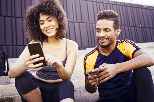 happy athletic latin woman and young black man looking the sports app in their mobile phones on the stairs during a rest in the workout, technology in sport and healthy lifestyle concept