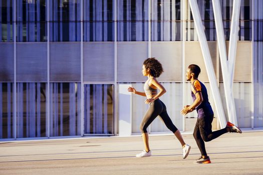athletic latin woman and happy black man running fast together at sunset in the city street with a building in the background. urban sports workout in couple and healthy lifestyle concept