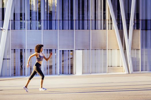 athletic latin woman running in the city street with a building in the background. urban sports workout in couple and healthy lifestyle concept
