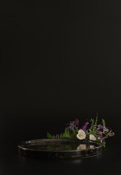 Black podium on the black background with flowers. Podium for product, cosmetic presentation. Creative mock up. Pedestal or platform for beauty products. Minimalist design, vertical view.