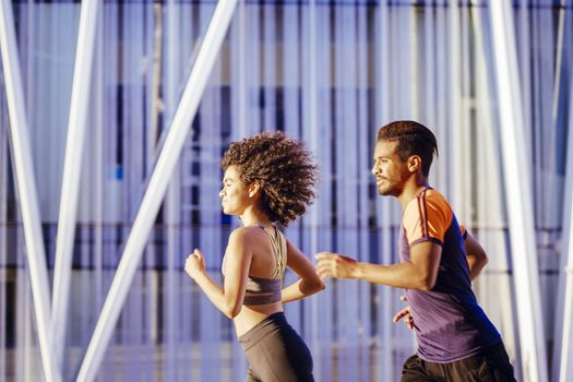 portrait of a beautiful latin woman and a sporty black man running together at sunset in the city street with buildings in the background. urban sports workout in couple and healthy lifestyle concept