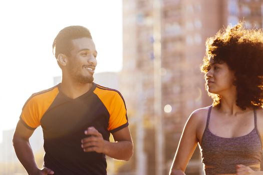 portrait of a happy black man running with a latin woman at sunset in the city street with buildings in the background. urban sports workout in couple and healthy lifestyle concept. sun flare effect