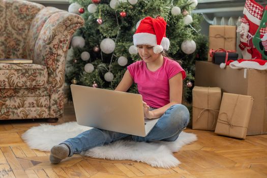 winter holidays, technology and children concept - little student girl with laptop computer at home over christmas tree background