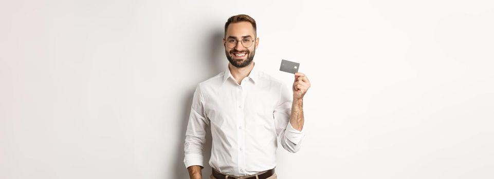 Handsome businessman showing his credit card, looking satisfied, standing over white background.