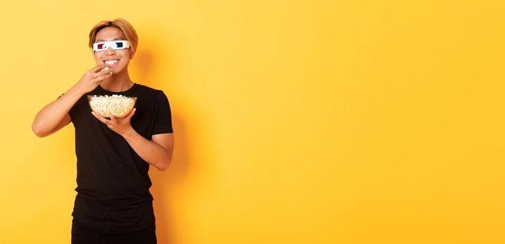 Joyful handsome asian guy with blond hair, watching movie or tv series in 3d glasses, eating popcorn and smiling amused, standing yellow background.