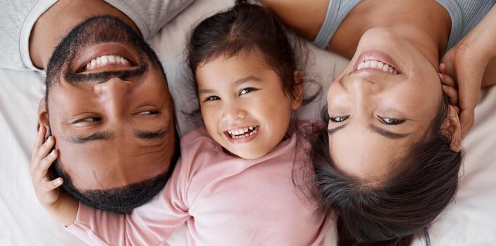Happy family with child on bed in a face portrait for interracial love, care and happiness together. Girl kid from Mexico with mother and father or parents relaxing at home and bonding from above.