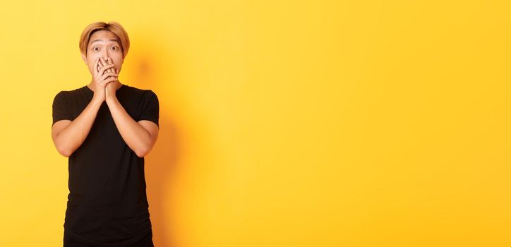 Portrait of surprised asian handsome guy with blond hair, cover mouth with hands and stare at camera impressed, yellow background.